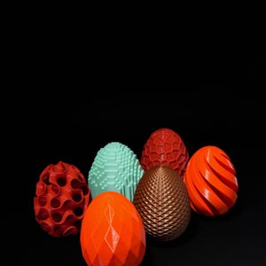 May include: A collection of six decorative, geometric-patterned eggs in various colors, including red, orange, brown, and teal, against a black background. The eggs feature diverse textures and designs, showcasing a modern aesthetic.