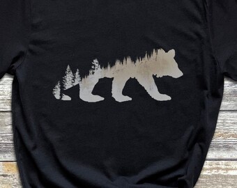 Può includere: Maglietta nera con una silhouette bianca di un orso con alberi all'interno del contorno dell'orso.