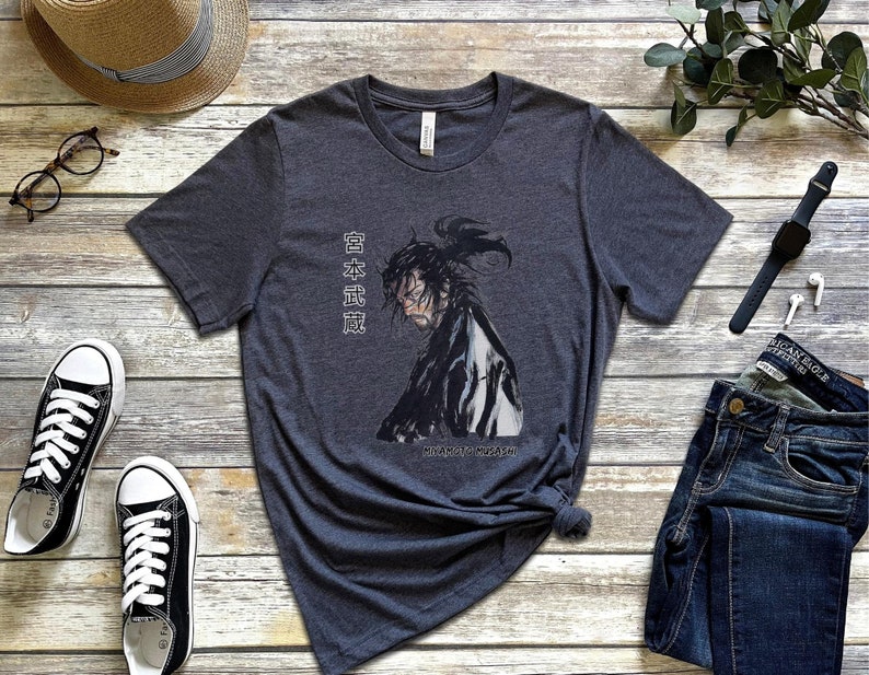 May include: A dark heather grey t-shirt with a black and white illustration of a samurai warrior. The shirt has the Japanese text "日本武道" printed on the right side of the image. The text translates to "Japanese Martial Arts".