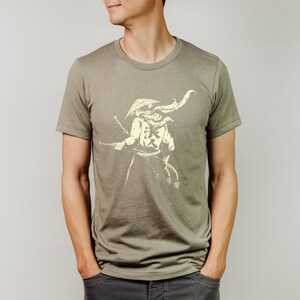 May include: Olive green t-shirt featuring a cream-colored illustration of a samurai warrior holding a katana. The samurai is depicted with a traditional helmet and attire. The shirt has a crew neck and short sleeves.