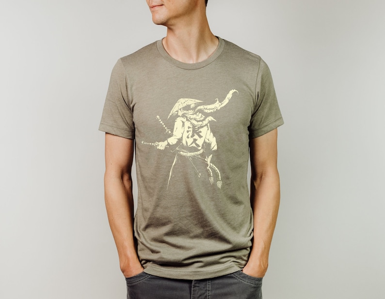May include: Olive green t-shirt featuring a cream-colored illustration of a samurai warrior holding a katana. The samurai is depicted with a traditional helmet and attire. The shirt has a crew neck and short sleeves.