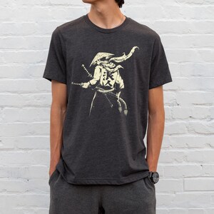 May include: Dark gray t-shirt featuring a cream-colored illustration of a samurai warrior with two swords and Japanese characters. The warrior is wearing a hat and a flowing scarf. The shirt is a crew neck style.