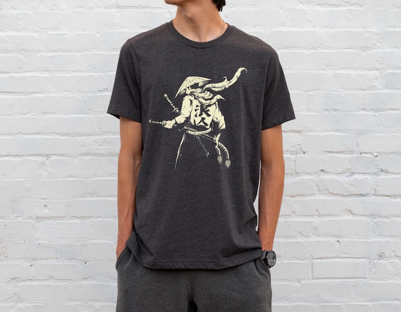 May include: Dark gray t-shirt featuring a cream-colored illustration of a samurai warrior with two swords and Japanese characters. The warrior is wearing a hat and a flowing scarf. The shirt is a crew neck style.