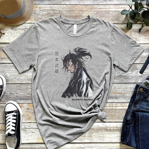 May include: A gray t-shirt with a black and white illustration of a samurai warrior. The text "Miyamoto Musashi" and Japanese characters are printed on the shirt.