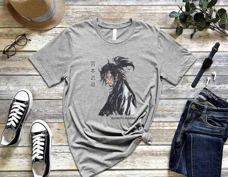 May include: A gray t-shirt with a black and white illustration of a samurai warrior. The text "Miyamoto Musashi" and Japanese characters are printed on the shirt.