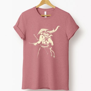 May include: A dusty rose t-shirt with a cream-colored illustration of a samurai warrior holding two swords. The samurai is wearing a helmet and armor, with Japanese characters on the chest. The shirt is hanging on a wooden hanger.
