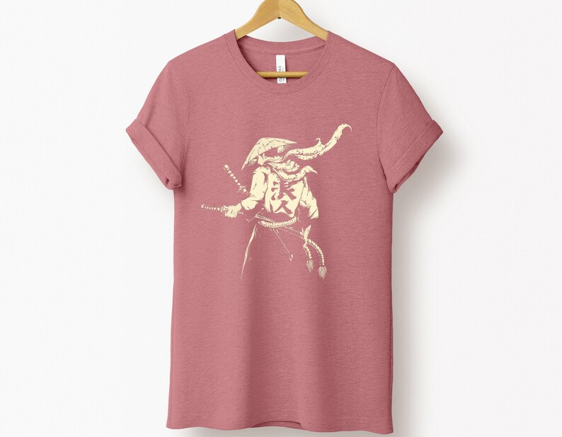 May include: A dusty rose t-shirt with a cream-colored illustration of a samurai warrior holding two swords. The samurai is wearing a helmet and armor, with Japanese characters on the chest. The shirt is hanging on a wooden hanger.