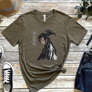May include: A green t-shirt with a black and white illustration of a samurai warrior. The text "日本武道" is printed on the shirt. The text "MIYAMOTO MUSASHI" is printed below the illustration.