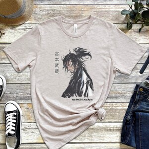 May include: A light beige t-shirt with a black and white illustration of a samurai warrior. The text "Miyamoto Musashi" and Japanese characters are printed on the shirt.