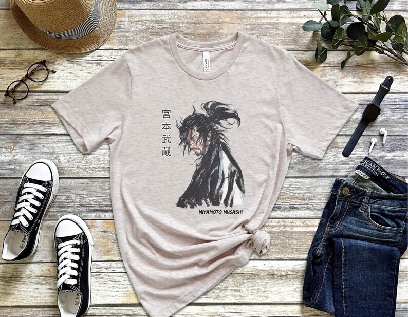 May include: A light beige t-shirt with a black and white illustration of a samurai warrior. The text "Miyamoto Musashi" and Japanese characters are printed on the shirt.