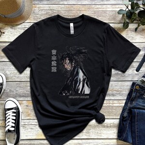 May include: Black t-shirt with a graphic of a person with long black hair and a white shirt. The text "日本武道" is printed on the shirt.