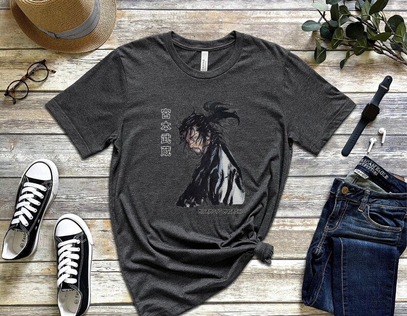 May include: A dark heather grey t-shirt with a black and white illustration of a samurai warrior. The Japanese text on the shirt reads "日本武道".