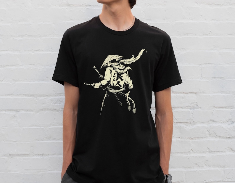 May include: Black t-shirt featuring a cream-colored illustration of a samurai warrior with two swords. The samurai is wearing a hat and armor, with Japanese characters on the back. The design is a detailed, stylized graphic.