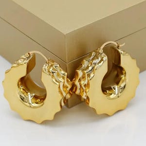 May include: A pair of gold-colored hoop earrings. The earrings have a unique, textured design with a scalloped edge. The earrings are displayed in front of a gold-colored box.