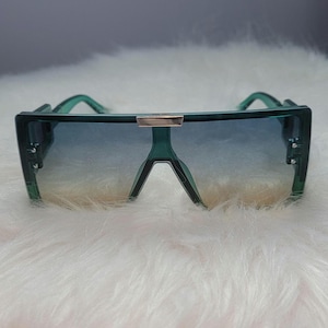 May include: Green oversized sunglasses with a gradient blue lens. The sunglasses have a rectangular frame with a silver bridge and a thick, angular design. The lenses transition from blue to a lighter shade.
