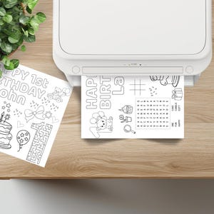 Puede incluir: Una impresora blanca imprime una p&aacute;gina para colorear en blanco y negro. La p&aacute;gina dice "HAPPY 1st BIRTHDAY John" e incluye im&aacute;genes de una tarta, un laberinto y un globo. Tambi&eacute;n se ve una segunda p&aacute;gina.
