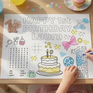 Puede incluir: Un mantel individual para colorear de cumplea&ntilde;os con el texto "HAPPY 1st BIRTHDAY Laura". El mantel individual presenta ilustraciones de una tarta, globos, un oso de peluche y un laberinto. Un ni&ntilde;o est&aacute; coloreando el n&uacute;mero uno con un cray&oacute;n azul.
