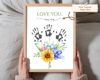 Puede incluir: Una impresión enmarcada blanca con un ramo de flores de acuarela y tres huellas de manos negras. El texto "LOVE YOU, nana" está escrito en verde en la parte superior de la impresión.