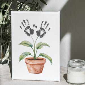 Puede incluir: Impresión en lienzo blanco con una ilustración de acuarela de una planta con flores de huellas de manos. La planta tiene hojas verdes y está en una maceta de terracota. Un tarro de cristal con una sustancia blanca está en primer plano.