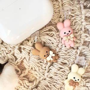 May include: Three small bunny charms with bows, in pink, brown, and white, attached to clear plastic clips. The charms are hanging from a white AirPod case.