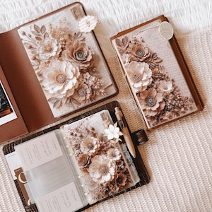 May include: Three brown leather planners and notebooks featuring floral cover art with beige and brown tones. The open planner displays clear plastic pockets, a thank you note, and a beige pen. Decorative paper clips are attached to the covers.