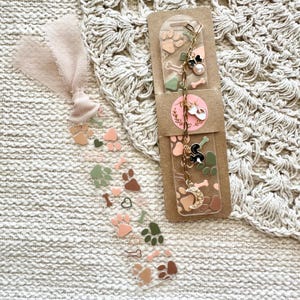 May include: A clear bookmark with a charm chain featuring paw prints, bones, a crescent moon, and a dog charm. The bookmark is decorated with paw prints, hearts, and bones in shades of pink, brown, and green.