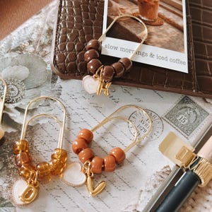 May include: Three gold-toned metal paperclip-style bookmarks adorned with beads and charms. One features dark brown beads with a heart and the letter H. Another has amber beads with a heart and the letter D. The third has terracotta beads with a heart and the letter V.