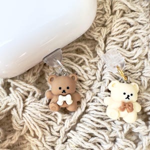 Könnte beinhalten: Zwei kleine Teddybär-Anhänger mit Schleifen, einer braun und einer weiß, befestigt an durchsichtigen Plastikclips. Die Anhänger hängen an einem weißen Hintergrund.
