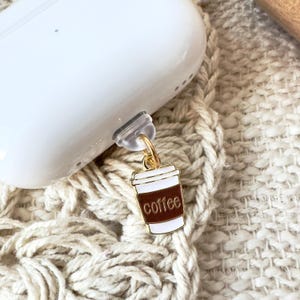 May include: A white Airpod case with a gold charm attached. The charm is a coffee cup with brown coffee and the word "coffee" written in white.