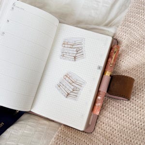 May include: Open notebook with grid paper, featuring two stickers of stacked books. A pink and gold pen with a brown leather pen holder rests on the right side. The notebook is open to a page with the date June 15.
