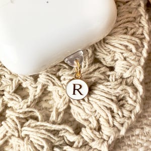 May include: White Airpod case with a gold charm attached. The charm is a small round disc with the letter "R" in gold lettering on a white background.
