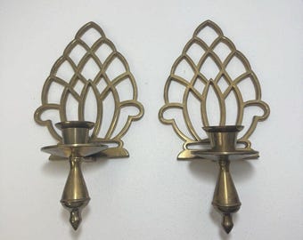 May include: Two brass wall sconces. Each sconce features a decorative openwork design with a candle holder. The sconces have a gold finish and are mounted on a white wall. The candle holders are designed to hold a standard size candle.