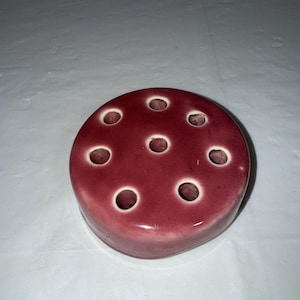 May include: A round, burgundy-colored ceramic flower frog with multiple holes. The flower arranging tool is designed to hold stems in place within a vase. The holes are evenly spaced across the top surface.