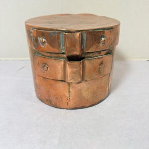 May include: A copper-colored, cylindrical container with a lid. The container has two bands around the body with rivets and a small opening. The metal has a patina, showing signs of age and use. The container is likely for storage.