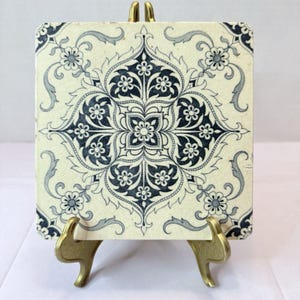 May include: A square ceramic tile featuring a dark blue and cream-colored symmetrical floral pattern with intricate scrollwork and a visible crackle glaze finish, displayed upright on a gold-toned metal stand against a plain white background.