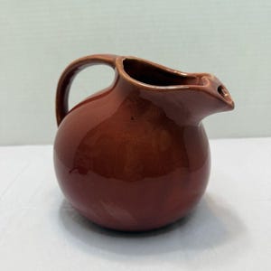 May include: A brown ceramic pitcher with a rounded body, a curved handle, and a spout. The pitcher has a glossy finish and is likely used for serving beverages. The overall shape is simple and functional.