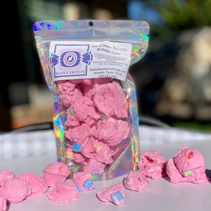 May include: A clear, iridescent bag filled with pink bubble gum ice cream scoops. The bag has a label with the text "Madi's Sweets" and "Ice Cream Scoops Bubble Gum." Several scoops are scattered in front of the bag.