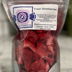 May include: A clear plastic bag filled with fresh, red strawberry slices. The bag has a label that reads "ADI'S SWEETS" and "Fresh Strawberries". The label also includes information about the product being made in a home kitchen and not inspected by the Department of State Health Services. It also includes the website and phone number for Madis Sweets And Treats.