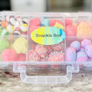 May include: A clear plastic box with a handle, divided into six compartments. Each compartment is filled with different pastel-colored candies. The box has a label that says "Snackle Box".