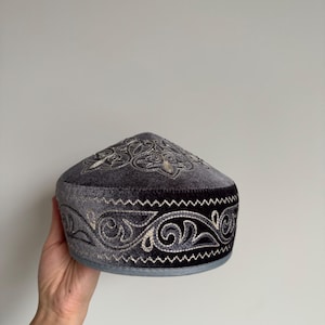 May include: A dark gray velvet cap with intricate silver embroidery. The cap features a rounded crown and a decorative band around the base. The embroidery showcases a swirling floral pattern, adding a touch of elegance to the headwear.