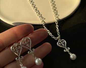 May include: A silver-toned jewelry set featuring a necklace and earrings. The necklace has a heart-shaped pendant with a dangling pearl. The earrings mirror the pendant design, also with pearl drops. The set is displayed on a white surface against a dark background.