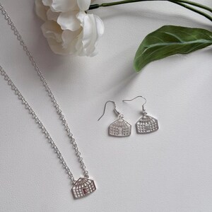 May include: A silver necklace and matching earrings set. The pendant and earrings feature a greenhouse design with a small red accent. The jewelry is displayed with white flowers and a green leaf.