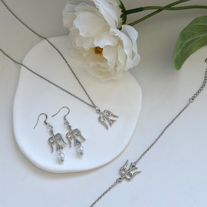 May include: A silver-toned jewelry set featuring a necklace, earrings, and bracelet. The necklace and earrings have a matching angel wing design, while the earrings also include a small pearl. The bracelet has a trident design. A white flower and green leaf are in the background.