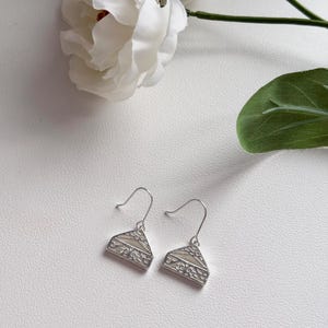 May include: A pair of silver-toned earrings with a triangular design. Each earring features a decorative pattern along the edges and a light-colored center. The earrings are displayed on a white surface, with a white flower and green leaf in the background.