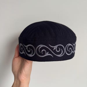 May include: A black cap with a decorative silver embroidered border. The cap is round with a slightly curved top and is held by a hand against a white background. The embroidered design features a repeating scroll pattern.