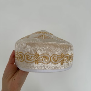 May include: A white velvet hat with a gold embroidered design around the brim.