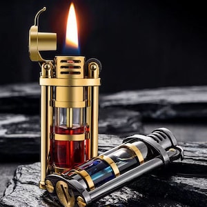 Op de afbeelding: Een gouden aansteker met een transparante glazen brandstofreservoir en een rode vloeistof erin. De aansteker heeft een vlam die uit de bovenkant komt. Er is een tweede aansteker op de afbeelding, die zwart en zilver is met een blauw en gouden ontwerp.