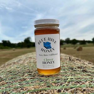 May include: A glass jar of clover honey labeled "Blue Hive Honey" with the text "Pure, Raw, Unfiltered" and "Made in Sullivan, MO" and "NET WT. 16 oz. (473g)". The jar is sitting on a bale of hay in a field.
