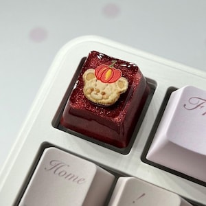 May include: A custom keyboard keycap featuring a bear-shaped cookie with a pumpkin on top. The keycap is a deep red color with glitter, and is surrounded by other light purple and white keys. The keyboard is on a white surface with pink polka dots.