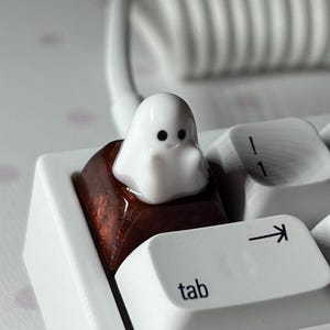 May include: A white ghost-shaped keycap sits on a brown keycap with a white keyboard in the background. The keyboard has keys labeled "tab", "capslock", and "enter".